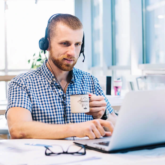 Smiling man wearing headphones and glasses typing on a keyboard at a desk with computer monitors displaying code